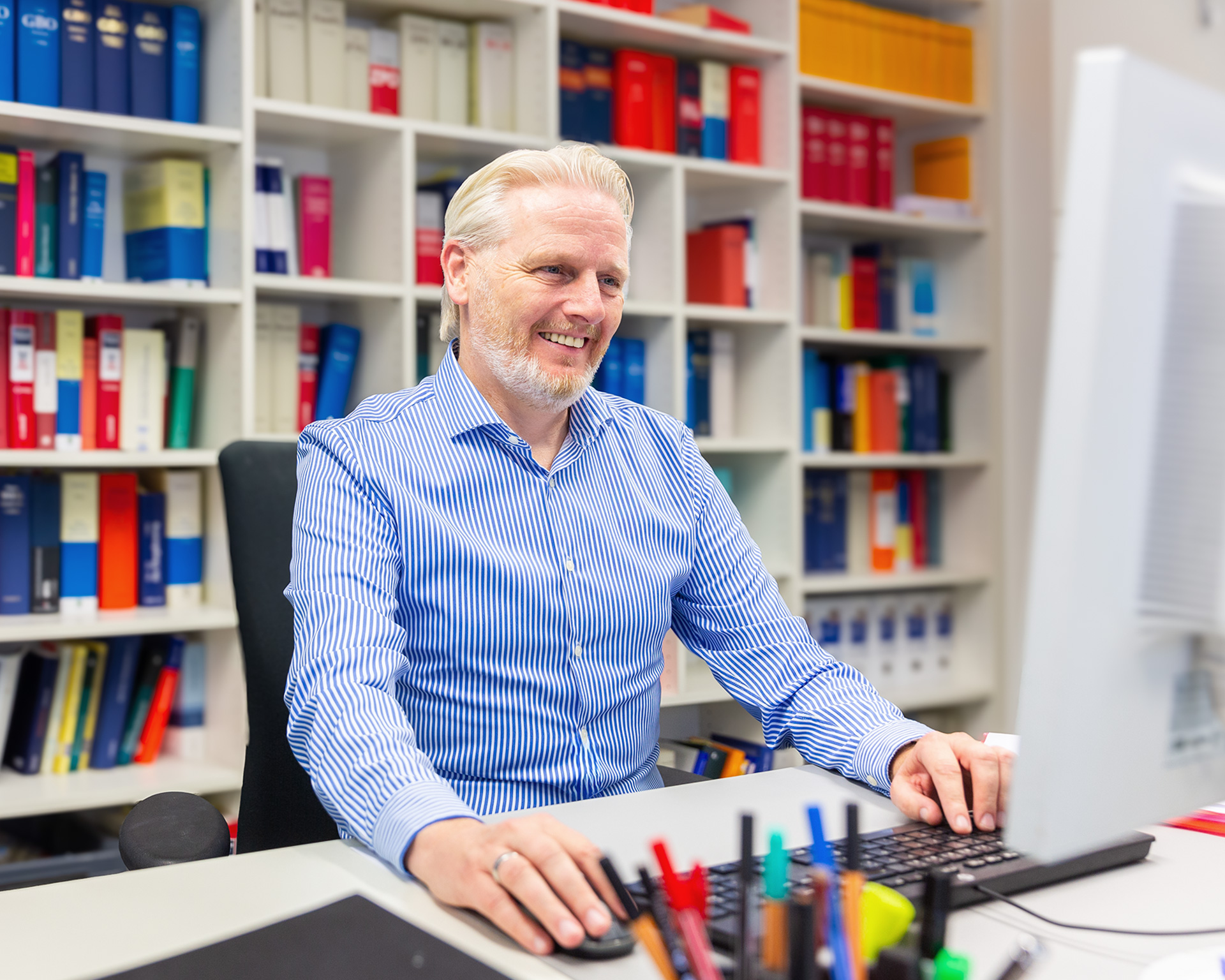 Notare-Wenz-Schueller-Office_DSC2313 Der Mitarbeitende Robert Müller sitzt an einem Computer, hinter ihm ist ein großes Bücherregal zu sehen